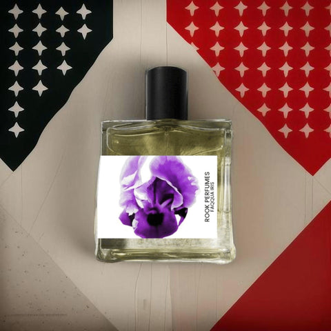 A square glass perfume bottle with a black cap and a label featuring a purple Faqqua Iris, the national flower of Palestine, is positioned against a background split into black and red triangular sections with white stars. The label reads "Faqqua Iris: A Scent for Gaza 30ml EDP RSX Rook Perfumes London | Unique Unisex Fragrance.